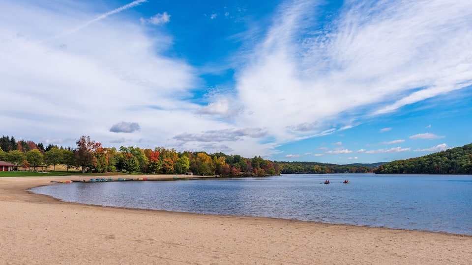 A lake with boats, a sandy beach, and autumn-colored trees.