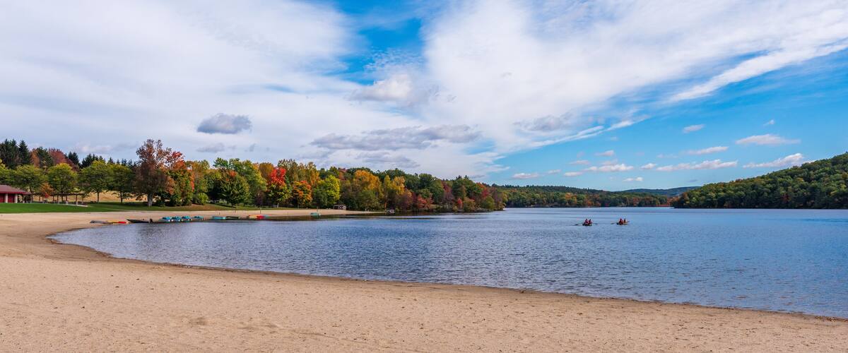 A lake with boats, a sandy beach, and autumn-colored trees.