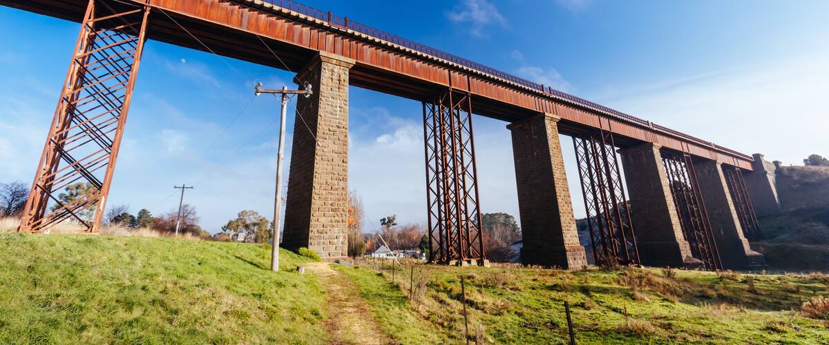 A large, rusted metal bridge with multiple supports, spanning over a grassy area with a clear blue sky.