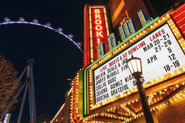 Une grande tente illuminée de néons, une grande roue en arrière-plan et une affiche indiquant les concerts et les événements de bowling à venir.