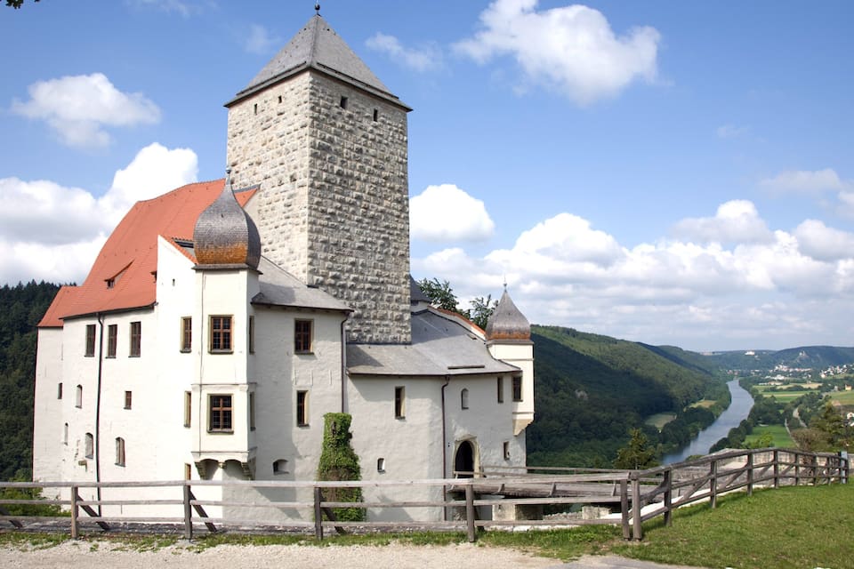 A castle with a red roof and a stone tower, surrounded by lush green hills and a river.