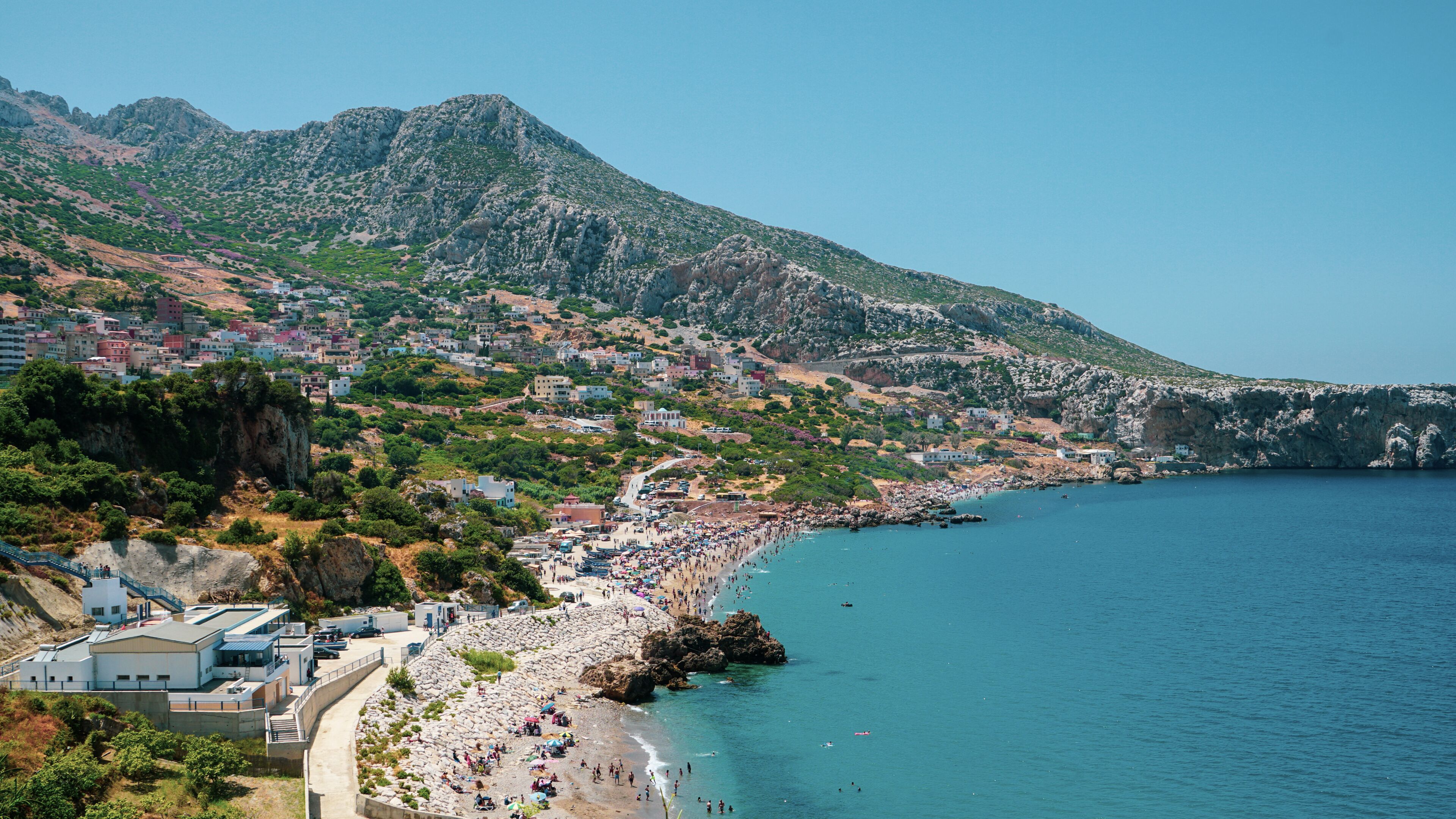 A coastal town with a beach, rocky cliffs, and a mountainous backdrop.