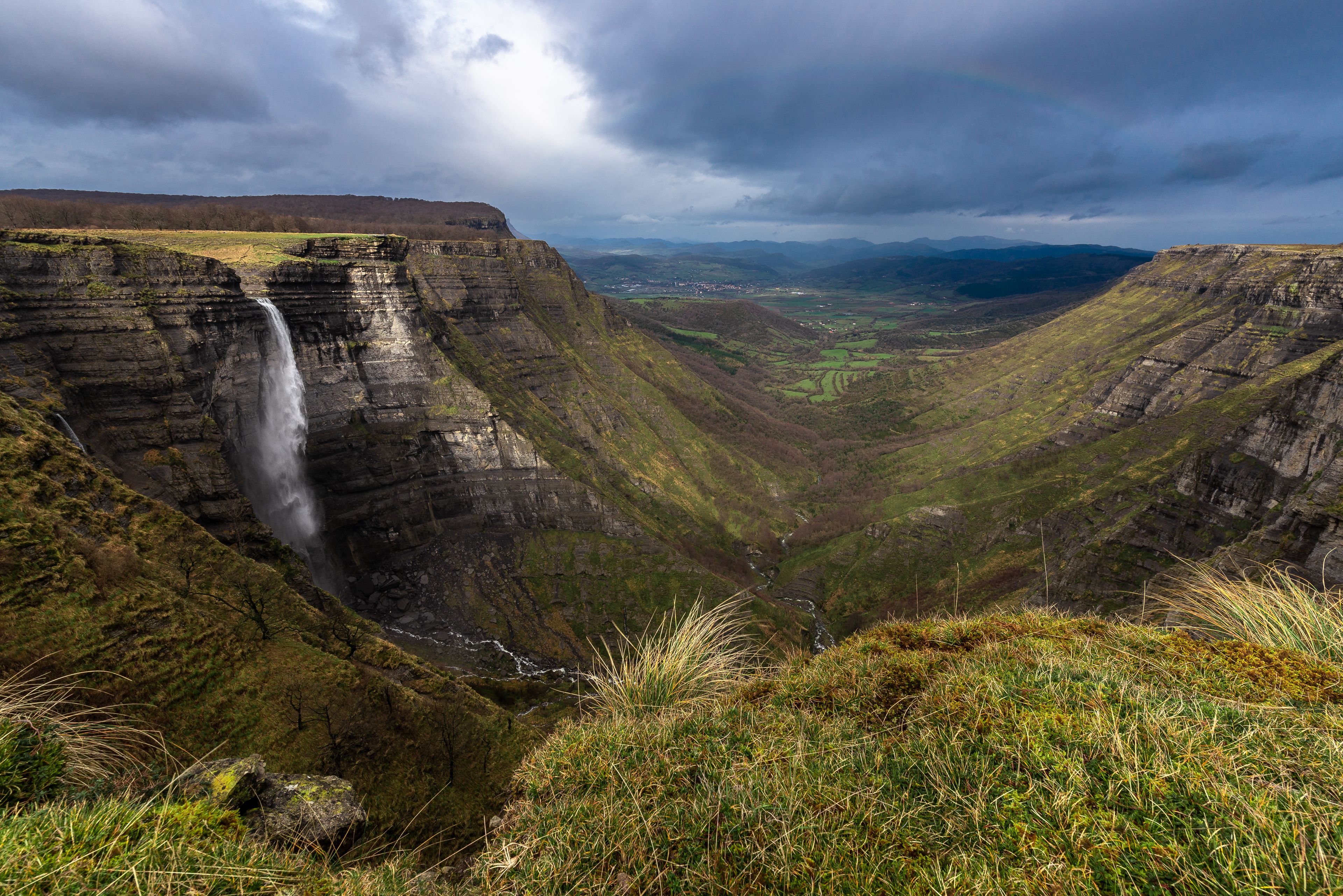 Una cascada se precipita por un acantilado escarpado, rodeada de colinas verdes exuberantes y un valle.