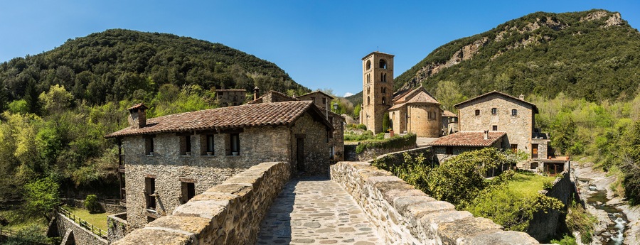 Un camino de piedra que conduce a un edificio histórico de piedra con una torre de campanas, rodeado de verdes colinas y un cielo azul despejado.