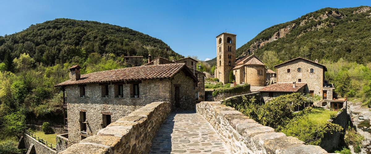 A stone pathway leading to a historic stone building with a bell tower, surrounded by lush green hills and a clear blue sky.