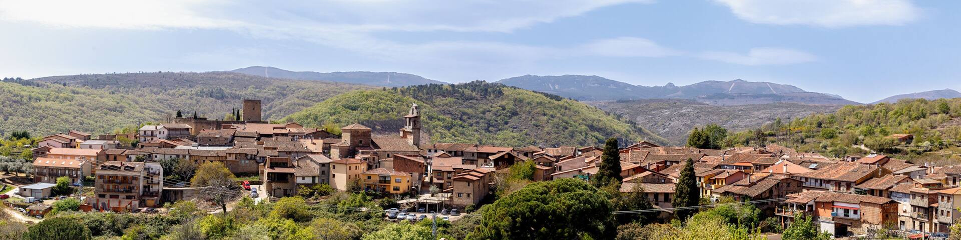 A village with terracotta-roofed buildings nestled in a valley, surrounded by lush green hills and mountains under a clear sky.