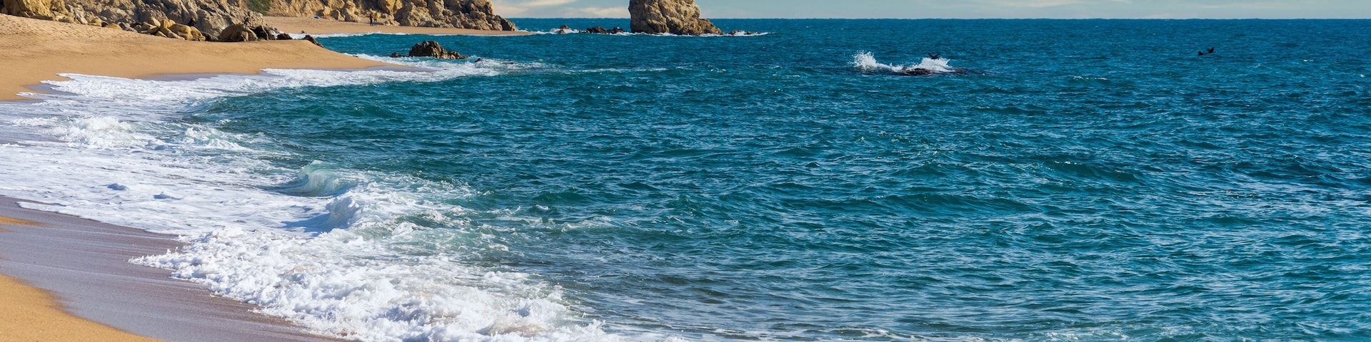 Uma praia arenosa com ondas, um afloramento rochoso e um céu azul límpido.