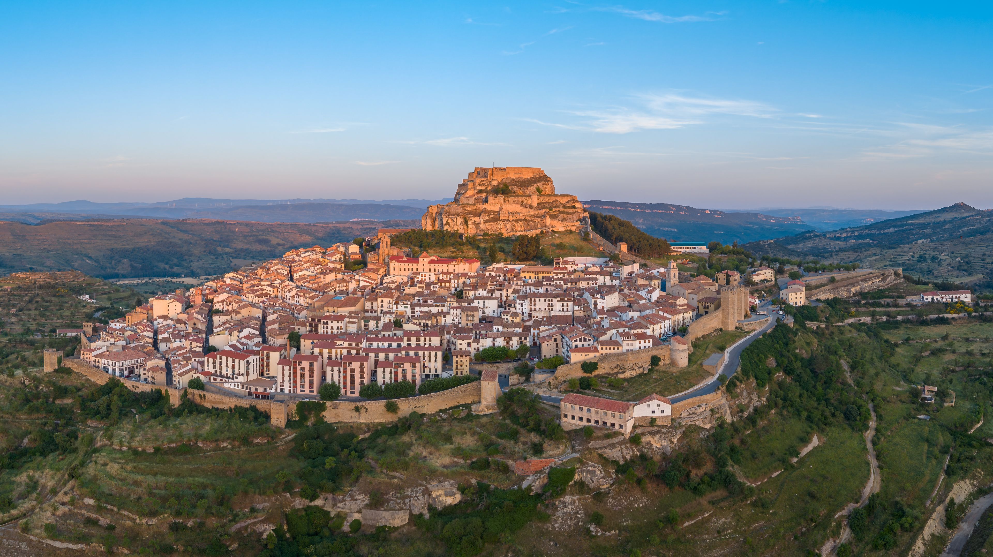 Panoramic aerial view of Morella, a medieval walled hilltop town in Els Ports, Castellon, Spain, featuring complete perimeter walls, towers, castle ruins, and surrounding valley at sunrise