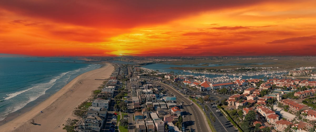 an aerial shot of the vast miles of homes and lush green trees around Huntington Harbour with blue water, majestic mountain ranges, cars driving on the street and powerful clouds at sunset