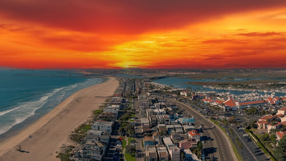 an aerial shot of the vast miles of homes and lush green trees around Huntington Harbour with blue water, majestic mountain ranges, cars driving on the street and powerful clouds at sunset