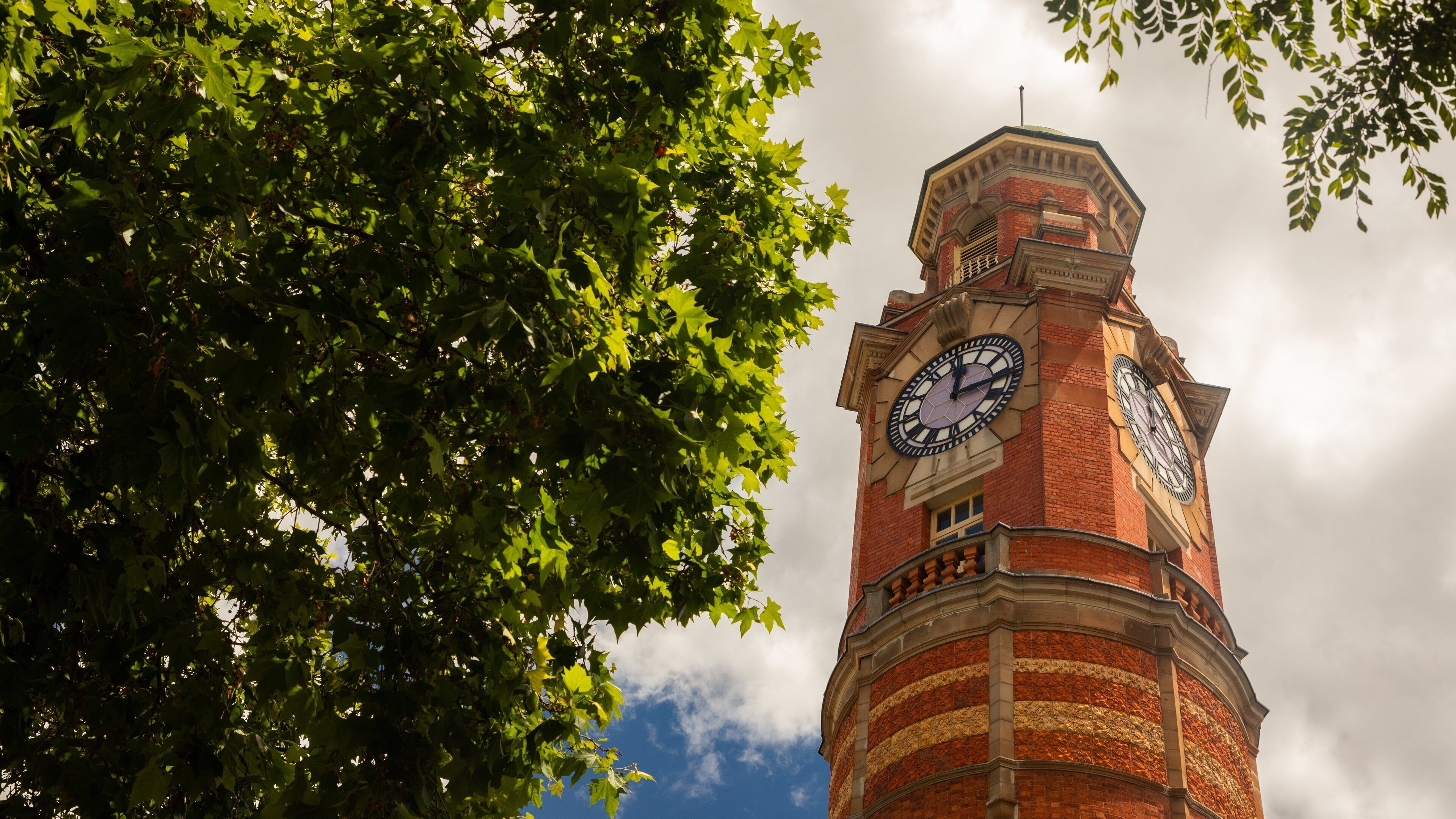 A clock tower with a brick facade and a large clock face, surrounded by lush green foliage.