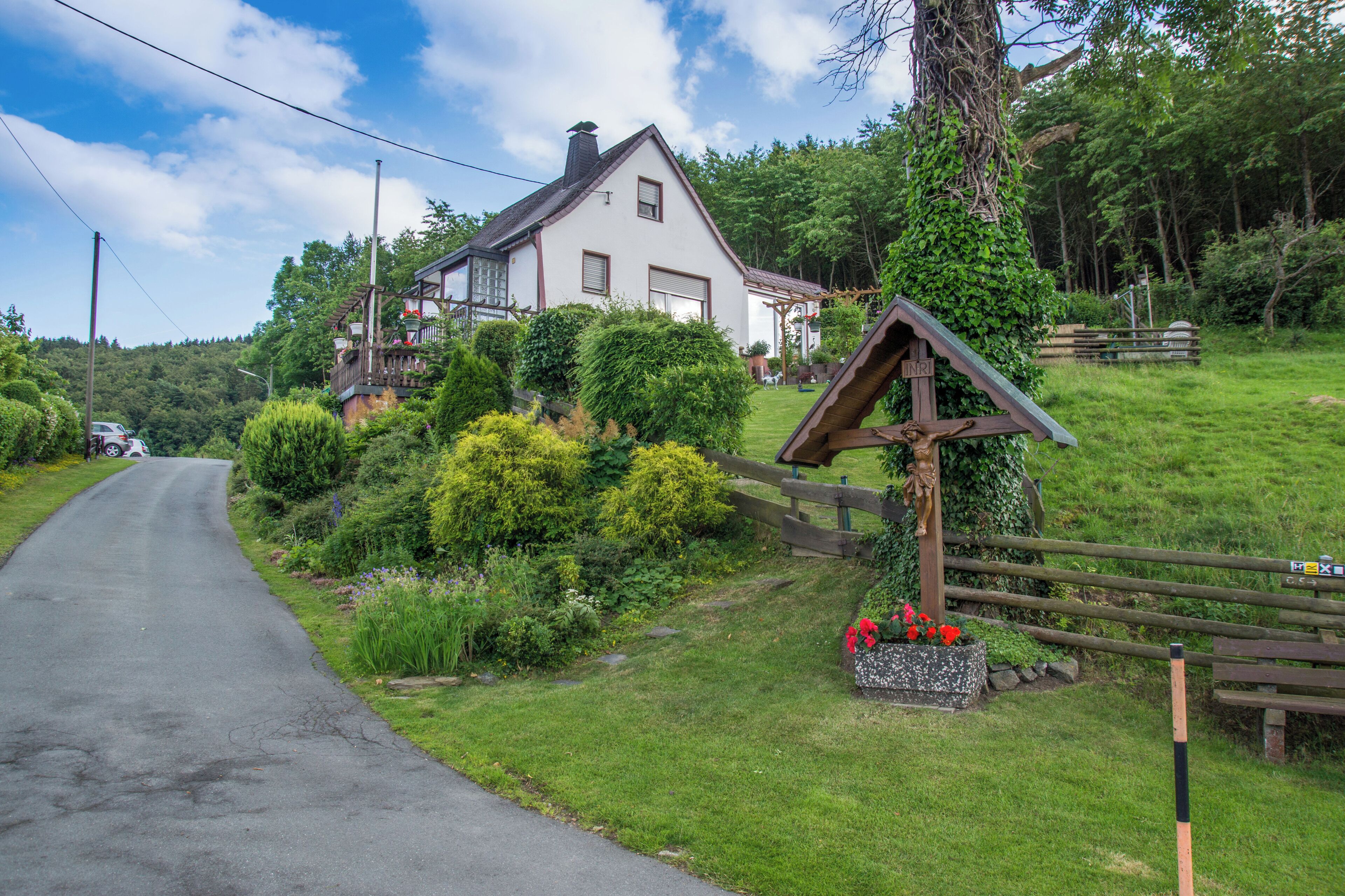 A rural road with a white house on a hill, surrounded by greenery and a wooden fence.