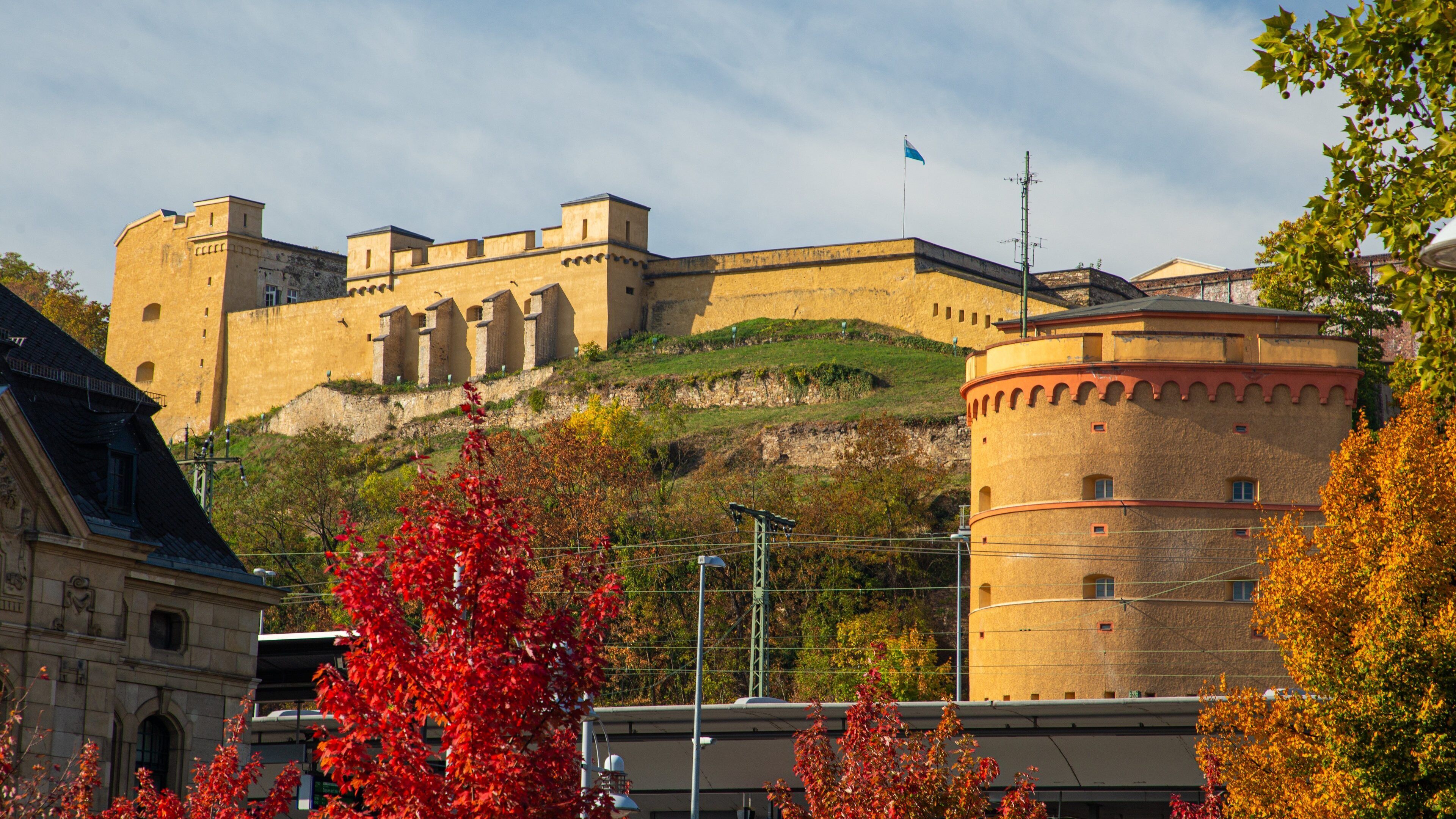 A historic castle with a prominent tower, surrounded by autumn foliage and modern buildings.