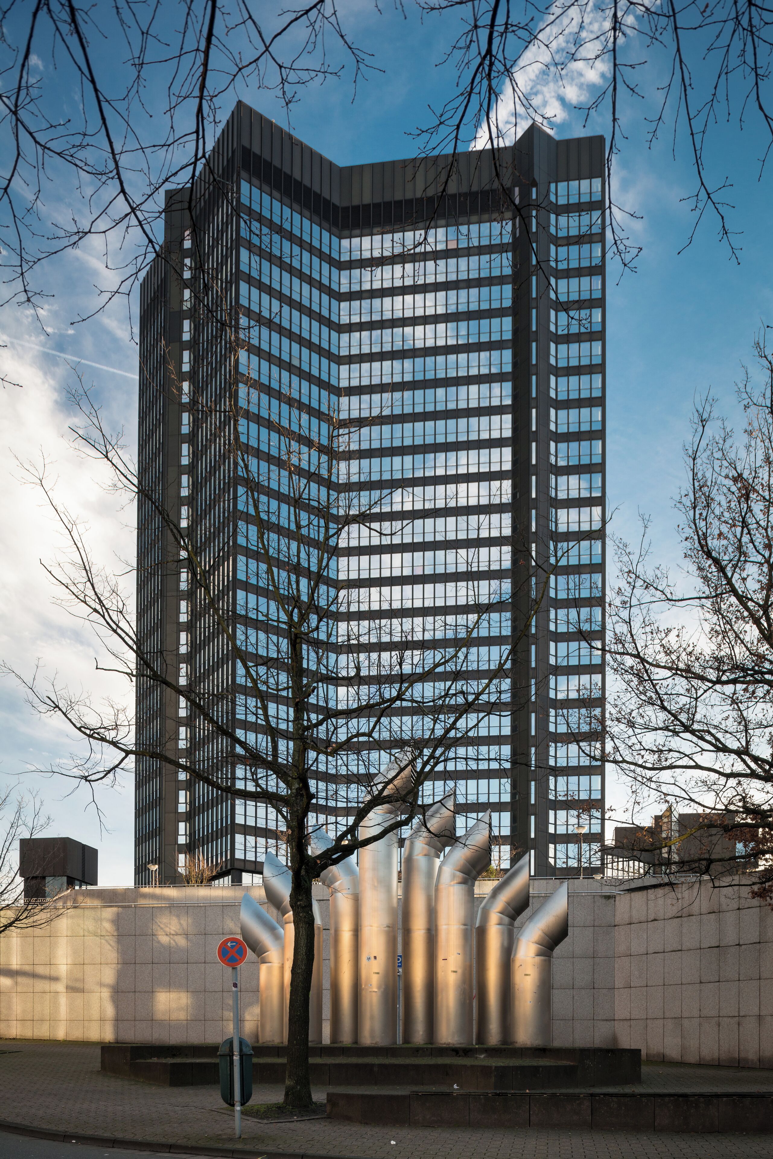 A tall modern building with a glass facade, surrounded by a paved area and leafless trees.