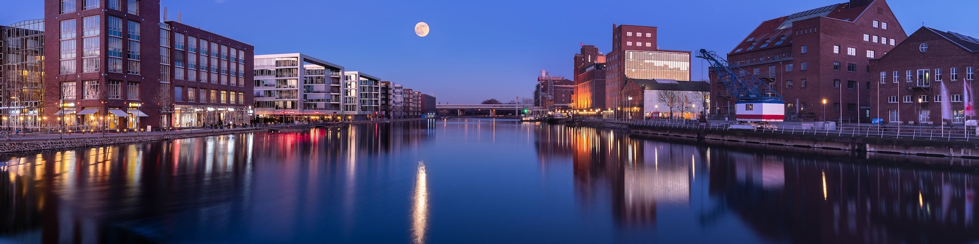 Ein ruhiger Fluss spiegelt Gebäude, einen Vollmond und eine Brücke in der Ferne wider.
