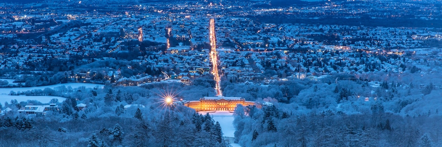 A snow-covered cityscape at dusk with a prominent illuminated tower and a large building with a snow-covered roof.