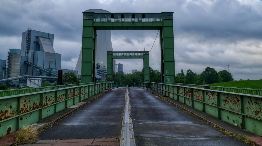 Eine grüne Metallbrücke mit Graffiti, die zu einer Stadtlandschaft mit modernen Gebäuden und einem bewölkten Himmel führt.