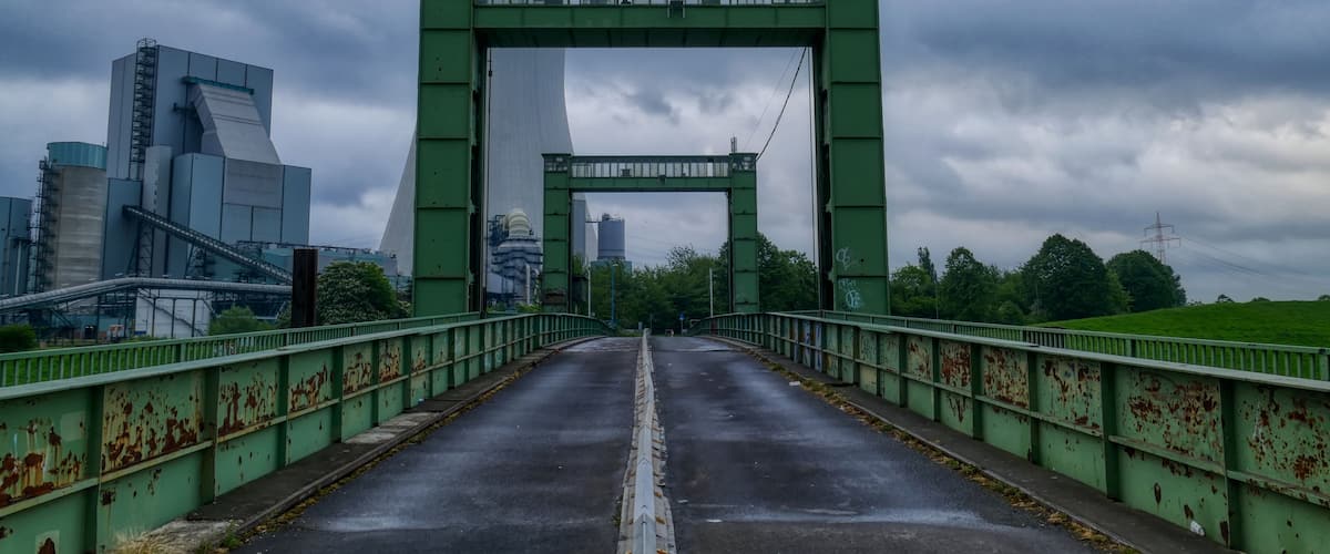 Eine grüne Metallbrücke mit Graffiti, die zu einer Stadtlandschaft mit modernen Gebäuden und einem bewölkten Himmel führt.