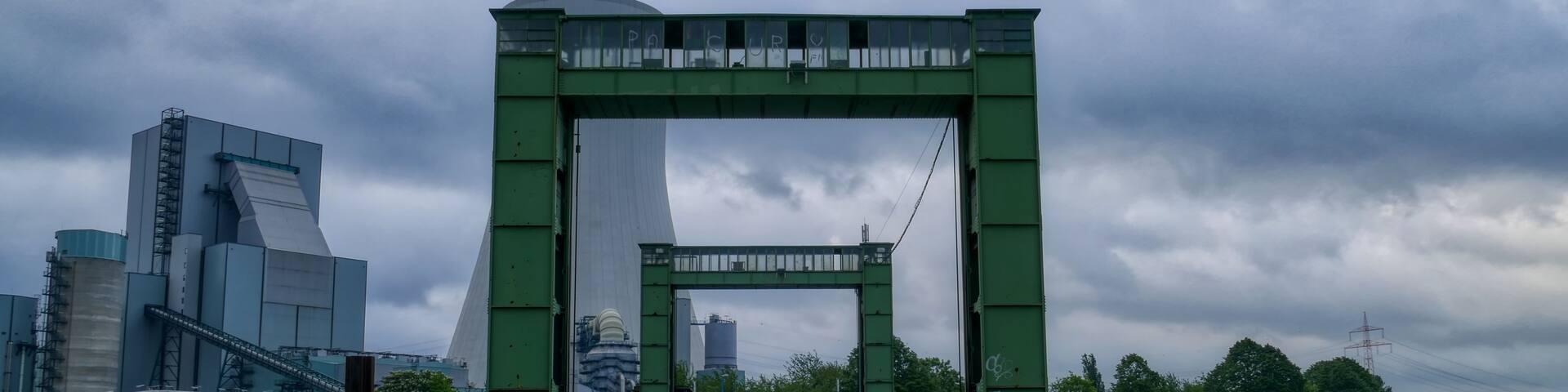 A green metal bridge with graffiti, leading to a cityscape with modern buildings and a cloudy sky.