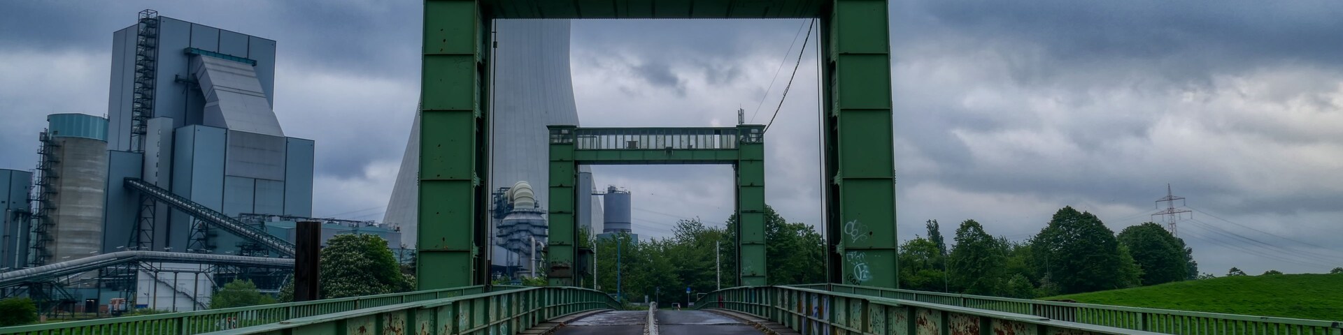 Uma ponte de metal verde com grafites, que leva a uma paisagem urbana com edifícios modernos e um céu nublado.