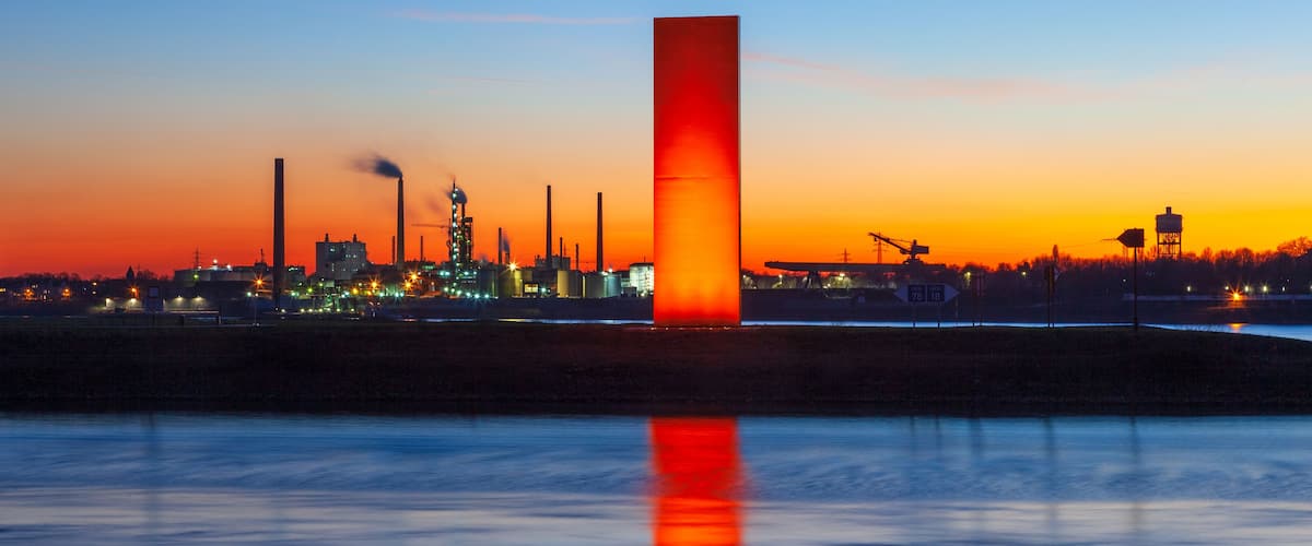 Ein Sonnenuntergang über einem Industriegebiet, mit einem markanten roten Turm, dessen Spiegelung sich im Wasser zeigt.