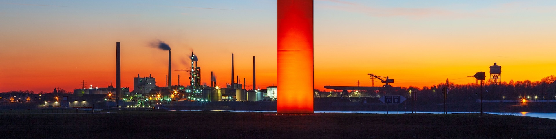Um pôr do sol sobre uma área industrial com uma torre vermelha imponente refletida na água.