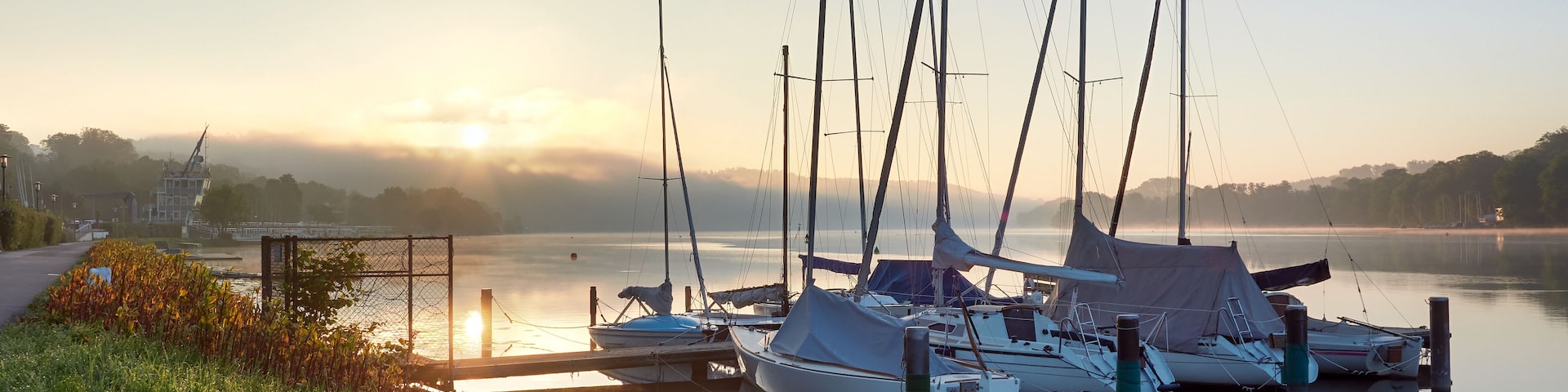 Un lac paisible où des voiliers sont amarrés au quai, entouré de verdure et baigné par la brume d’un lever de soleil.