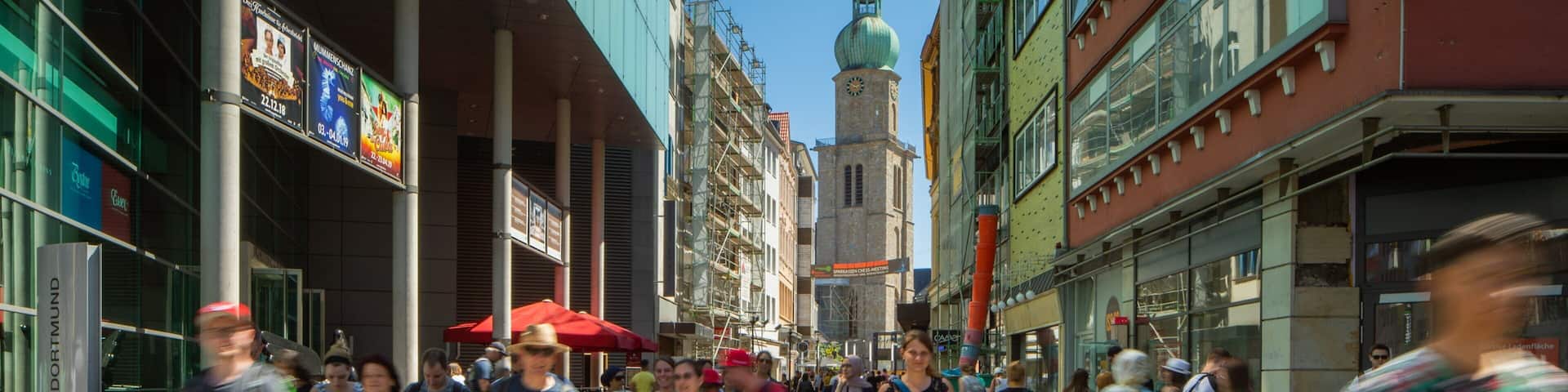 A busy urban street with pedestrians, a prominent church tower, and various commercial buildings.