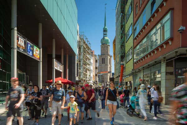 Une rue animée en plein centre-ville, avec des piétons, un clocher d’église imposant et divers bâtiments commerciaux.
