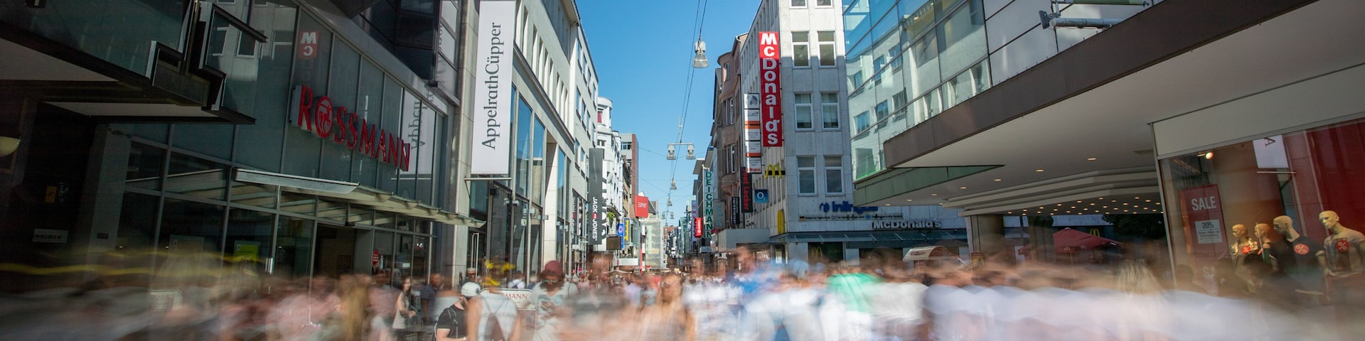 Uma rua urbana movimentada com lojas como Rossmann e McDonald's.