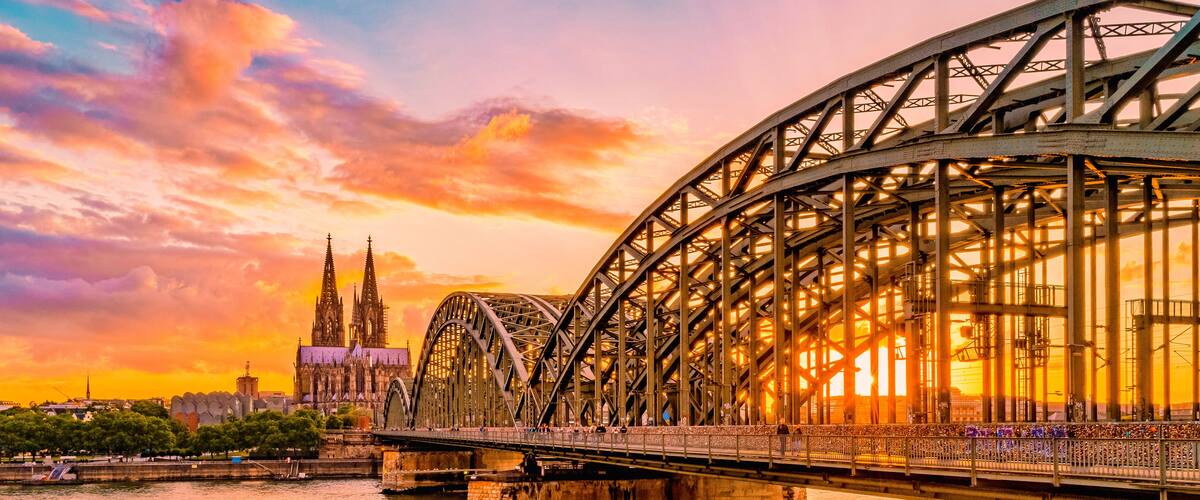 Eine Stahlbrücke überspannt einen Fluss, im Hintergrund ist eine Kathedrale bei Sonnenuntergang zu sehen.