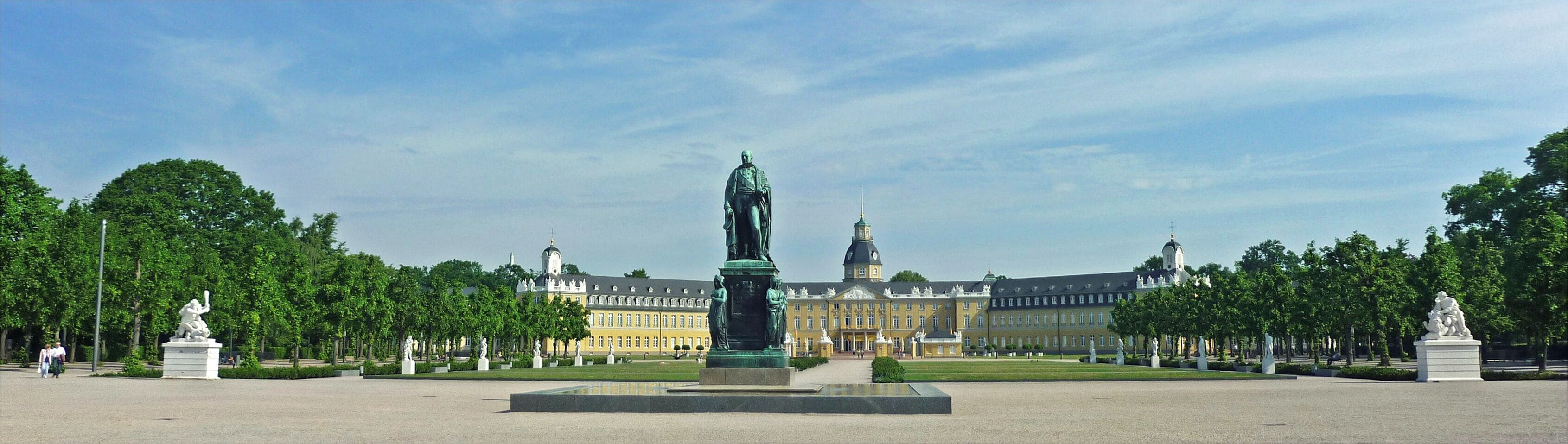 A large open square with a central statue, surrounded by trees and a grand building with multiple towers.