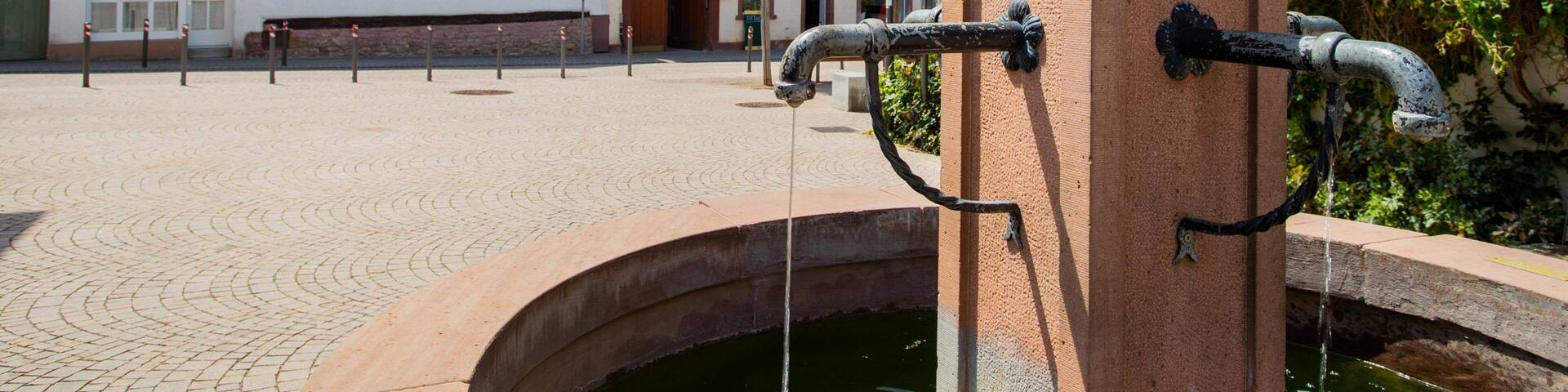 A traditional European town square with historic buildings, a water fountain, and cobblestone pavement.