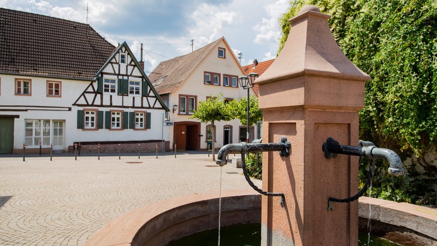 Ein traditioneller europäischer Stadtplatz mit historischen Gebäuden, einer Springbrunnen und Kopfsteinpflaster.