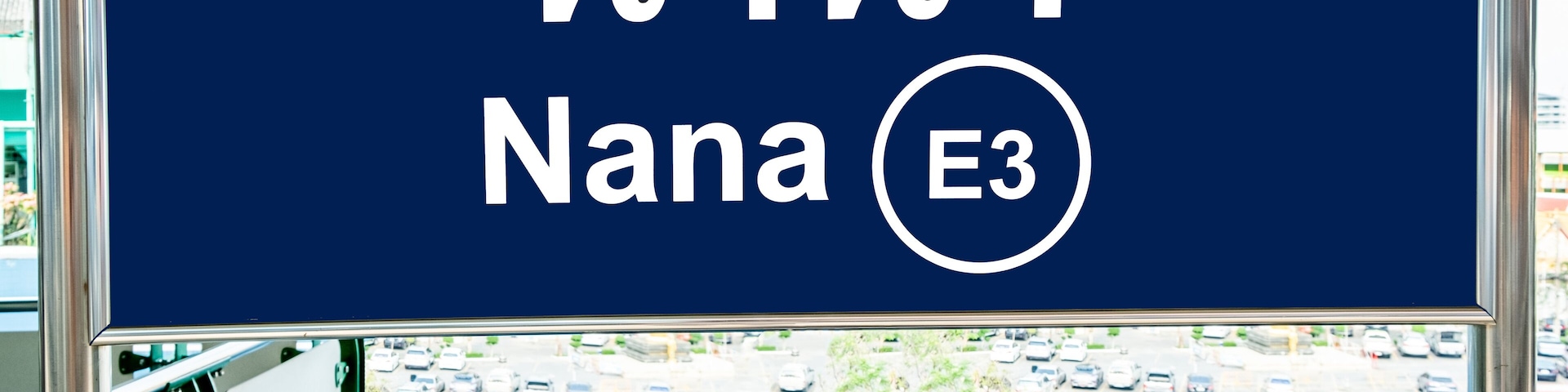 A blue sign with Thai text and a white circle, viewed from a window overlooking a parking lot.