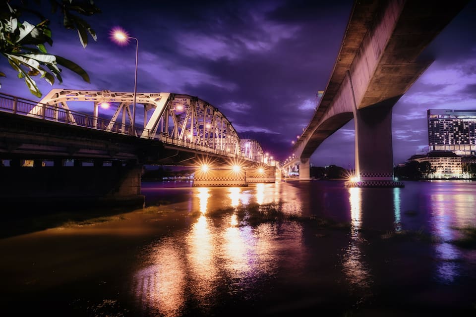 A nighttime view of a bridge with illuminated lights reflecting on the water, a large building with lights, and a purple sky.