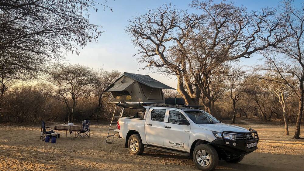 A white pickup truck with a roof rack and a tent set up in a dry, wooded area.