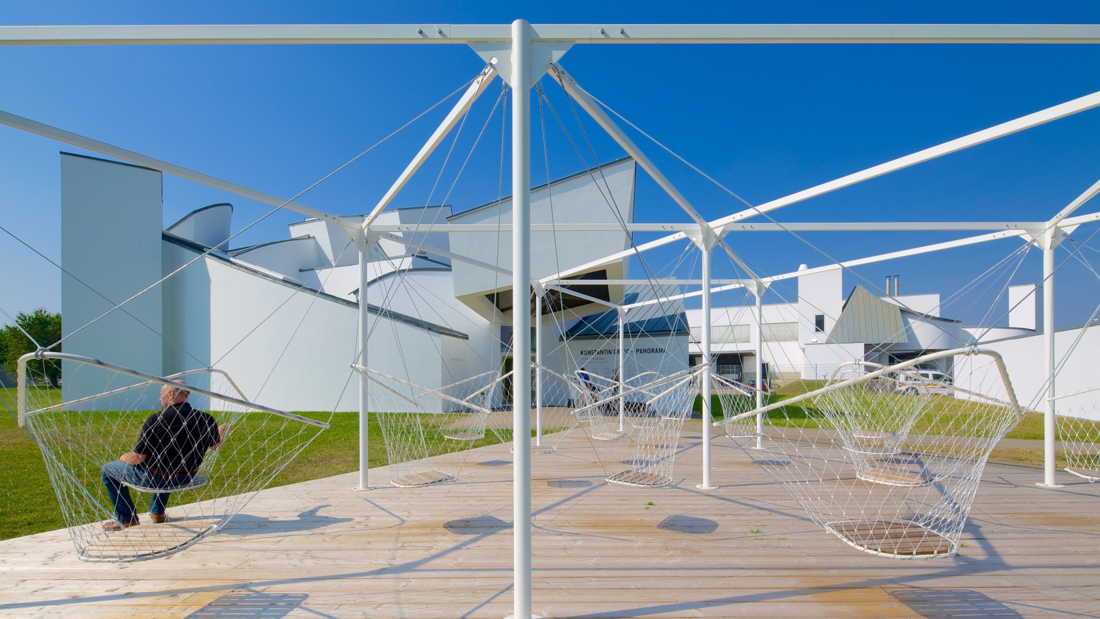 A modern architectural structure with a geometric design, featuring a large white building in the background and a person sitting on a swing-like chair in the foreground.
