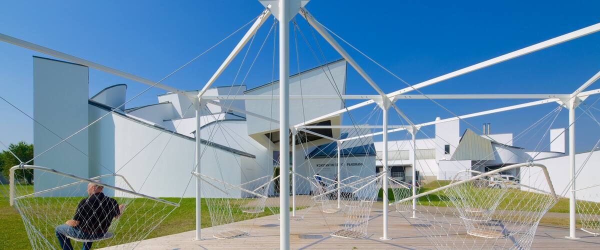 A modern architectural structure with a geometric design, featuring a large white building in the background and a person sitting on a swing-like chair in the foreground.