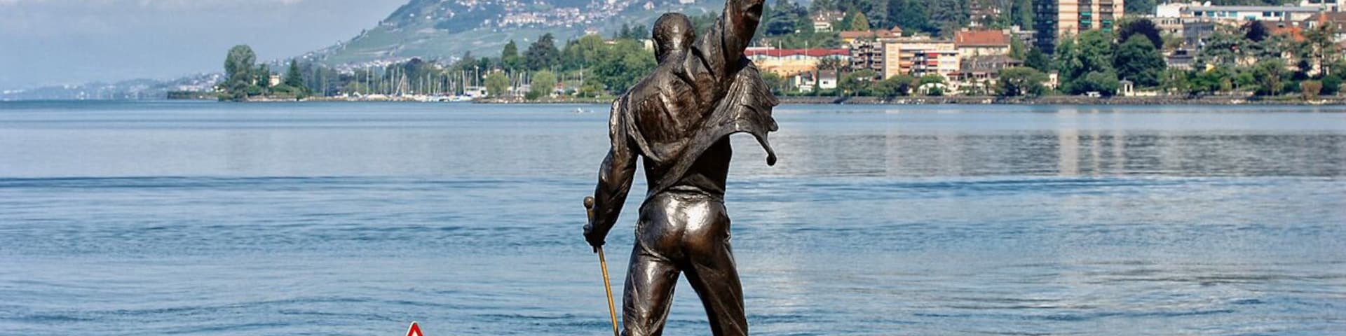 Estátua de homem com braço levantado à beira de um lago, com uma placa de aviso e uma bóia em primeiro plano.