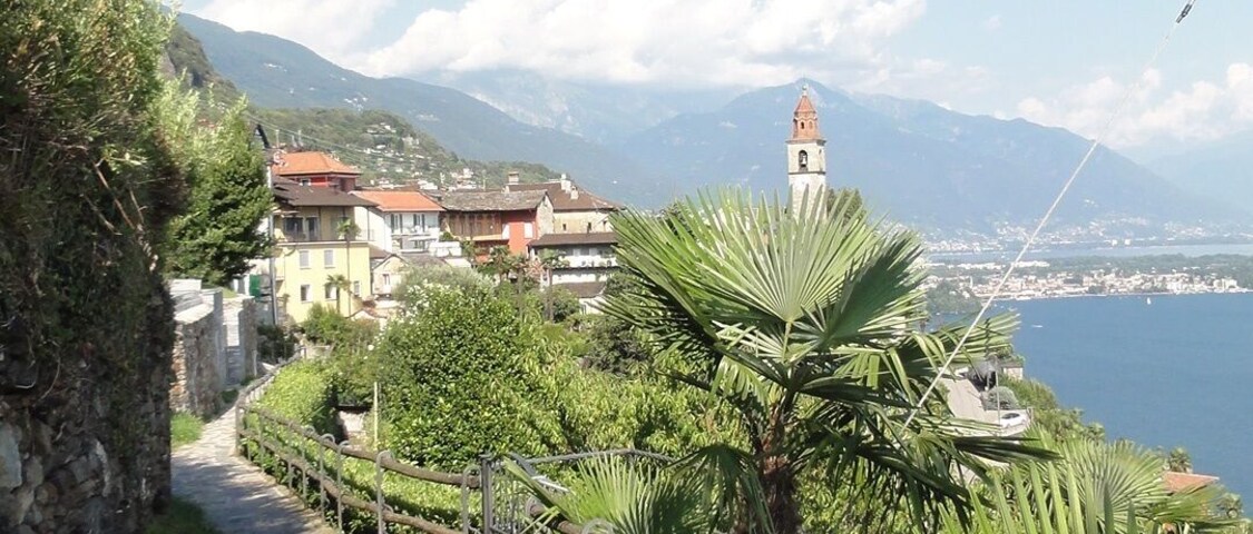 Uma trilha de pedra leva a uma vila costeira com uma torre de sino, cercada por vegetação exuberante e montanhas ao fundo.