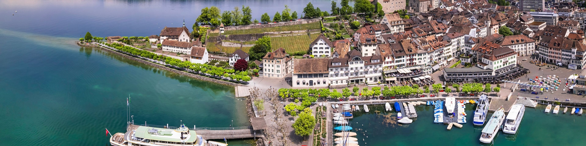 Eine Stadt am Seeufer mit historischen Gebäuden, einem Bootsanleger und einer Burg im Hintergrund.