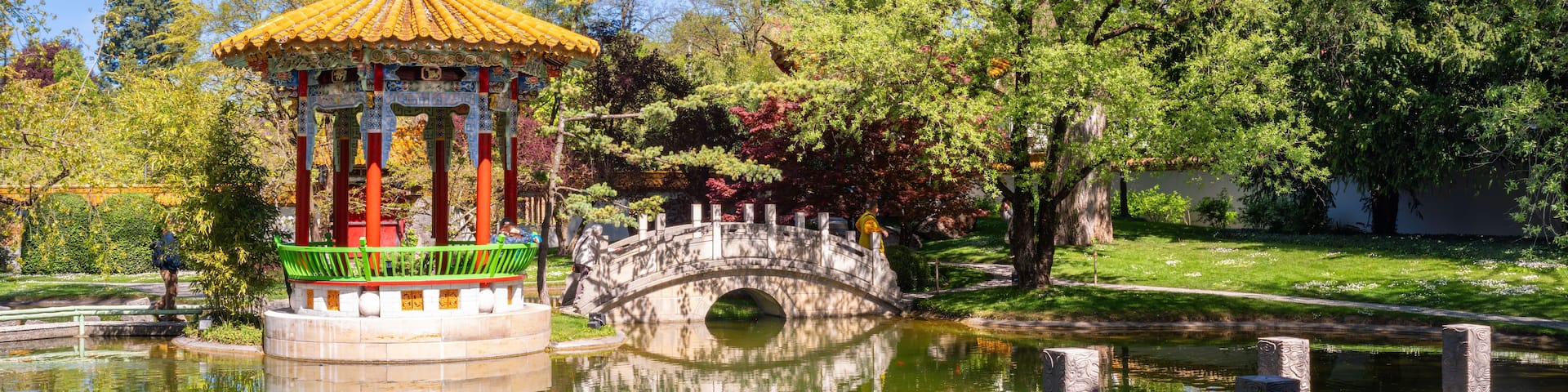A traditional pavilion with a tiled roof, a stone bridge over a pond, and lush greenery.