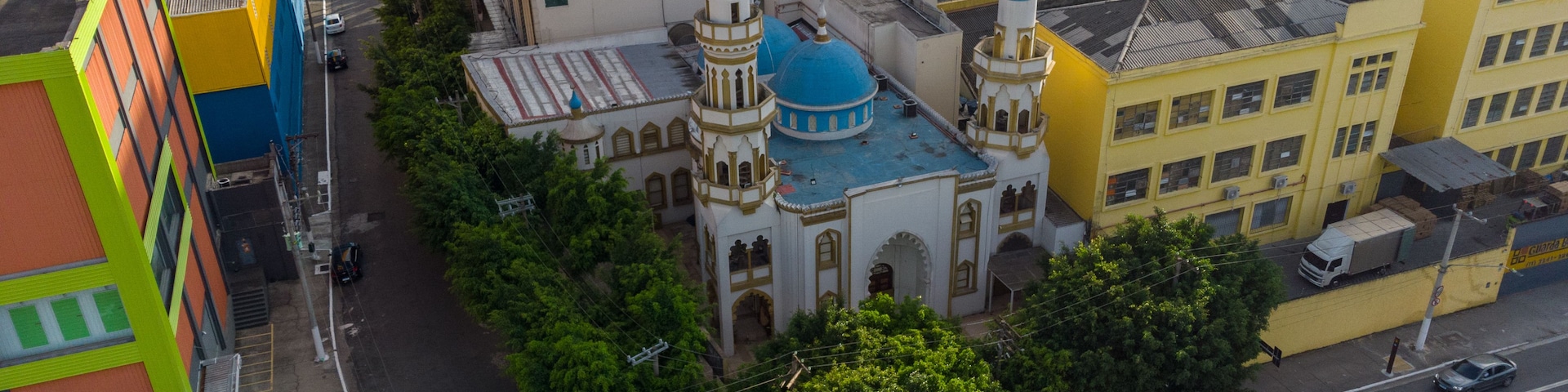 Un paysage urbain dominé par une mosquée imposante, avec ses coupoles bleues et son minaret, entourée de bâtiments aux couleurs vives et bordée d’une rue animée.
