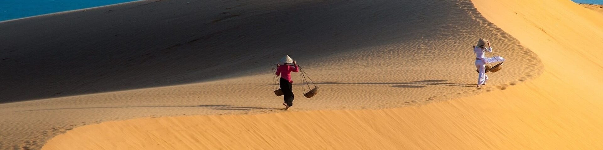 Two people walking on a sand dune with a body of water in the background.