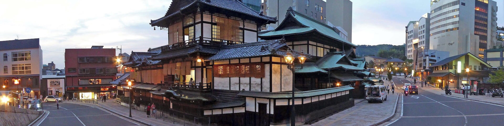 A traditional Japanese building with a sloped roof and wooden structure, located in an urban area with modern buildings nearby.