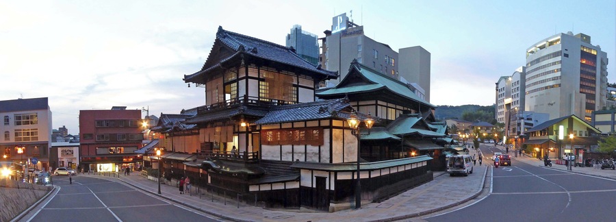 A traditional Japanese building with a sloped roof and wooden structure, located in an urban area with modern buildings nearby.