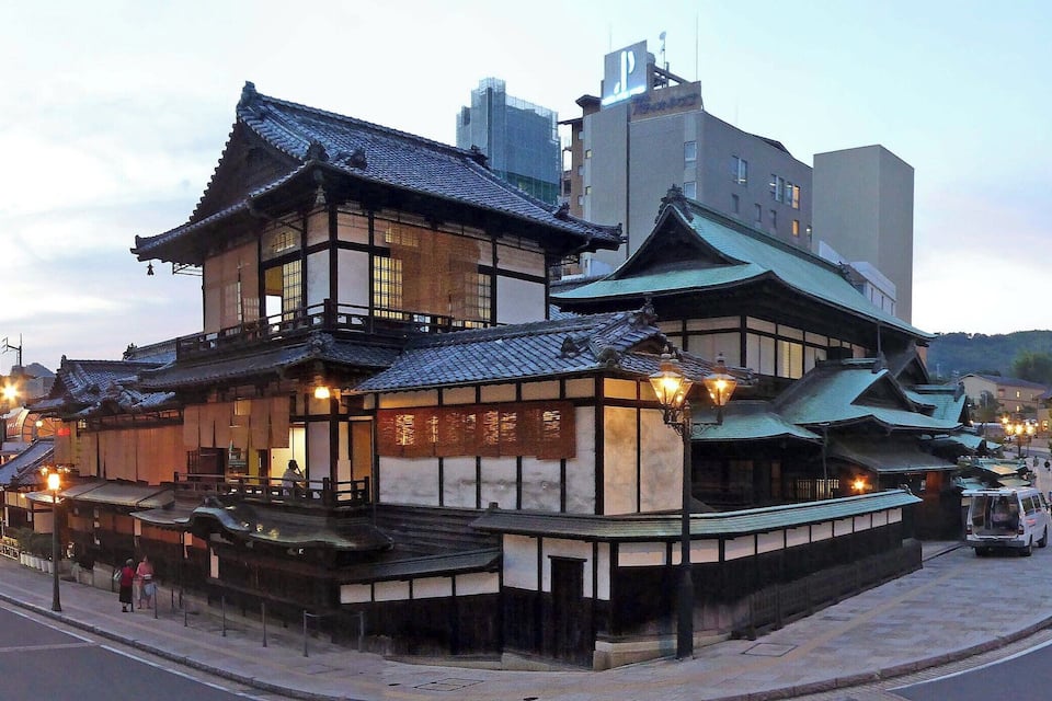 A traditional Japanese building with a sloped roof and wooden structure, located in an urban area with modern buildings nearby.