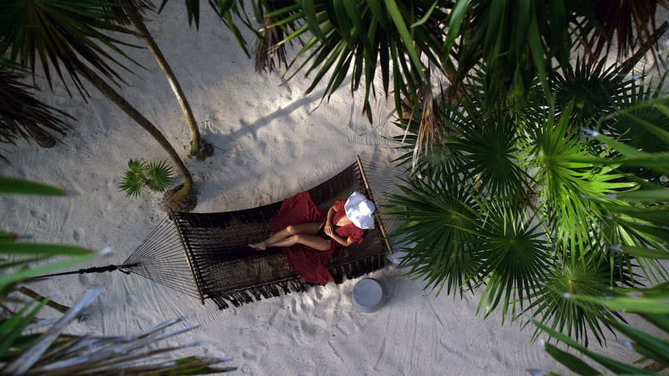 Woman rests in loose red summer dress and hat hanging in hammock surrounded by palm trees at sunset