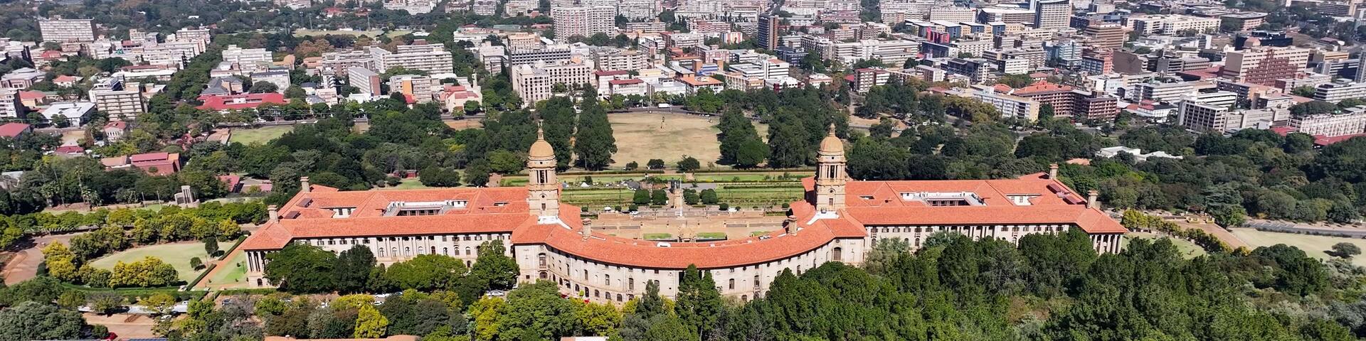A large historical building with a red roof, surrounded by dense greenery and a sprawling cityscape in the background.